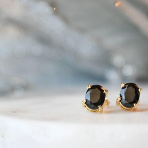 May include: A pair of gold stud earrings with oval-shaped black gemstones. The earrings are set on a white surface with a blurred, light-colored background. The gemstones have a polished, reflective surface.