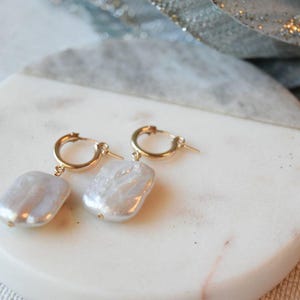 May include: A pair of gold hoop earrings with a large, off-white baroque pearl dangling from each hoop.