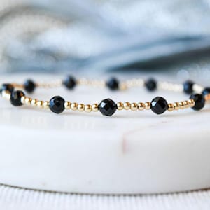 May include: A delicate gold bracelet with black gemstone beads. The bracelet is made with small gold beads and larger black beads.