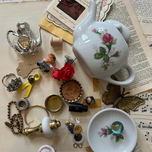 May include: A collection of assorted small objects including a white ceramic teapot with a pink rose pattern, a silver-tone metal teapot miniature, a white ceramic saucer with a rose motif, a brass butterfly, a blue marble, and various vintage trinkets.