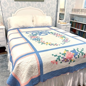May include: A white quilt featuring a central pixelated floral wreath design in shades of pink, blue, yellow, and green. The quilt has a blue and pink border with a scalloped edge and a quilted texture, draped over a bed with white pillows.