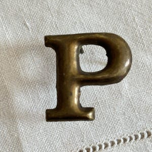 May include: A brass letter P with a smooth, metallic finish and a serif typeface design. The letter features a rounded top loop and a vertical stem with a flat base, showing a warm, aged golden tone with subtle variations in the metal surface.