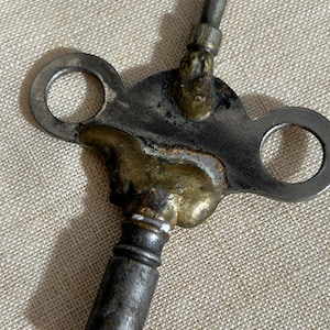 May include: A metal winding key featuring a T-shaped handle with two circular openings on either side. The central portion of the key is reinforced with a textured, brass-colored solder or metal buildup, connecting the handle to the cylindrical shaft.