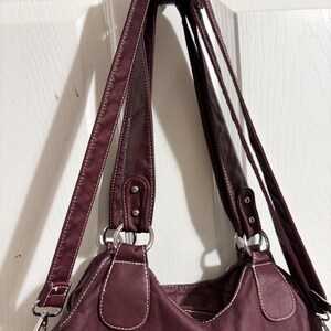 May include: A burgundy handbag with white stitching and silver-tone hardware. The bag has two shoulder straps and a crossbody strap. The bag is hanging on a black hook against a white door.