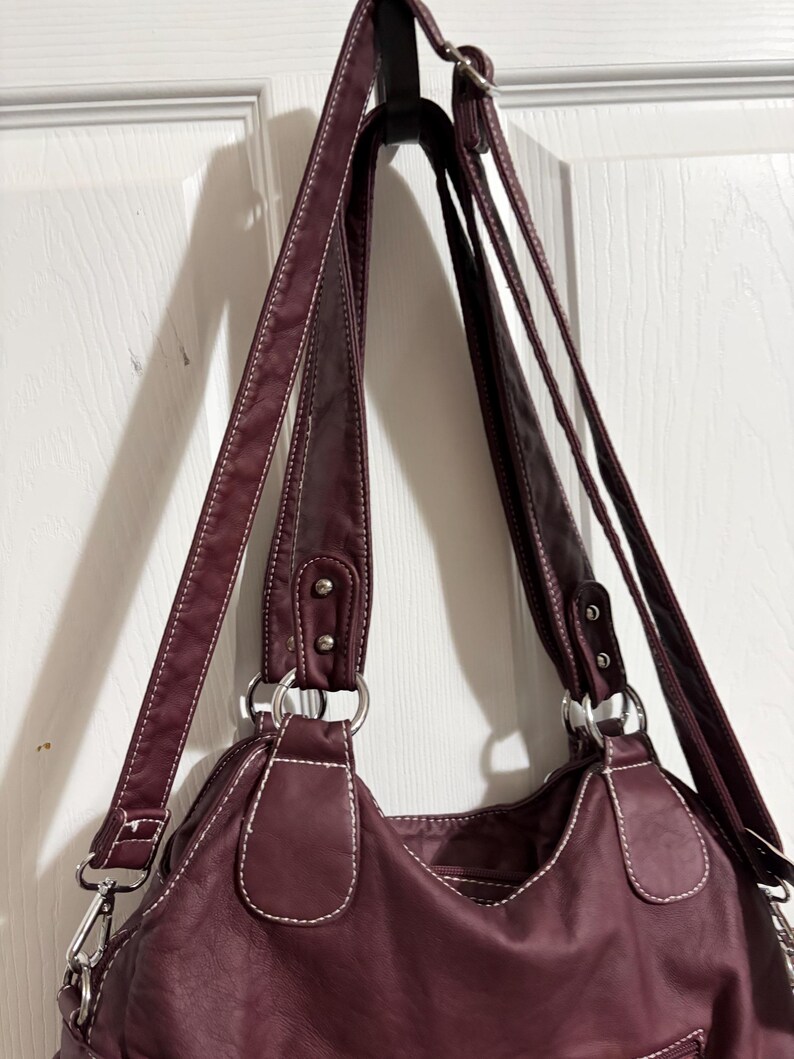 May include: A burgundy handbag with white stitching and silver-tone hardware. The bag has two shoulder straps and a crossbody strap. The bag is hanging on a black hook against a white door.