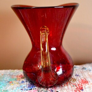 May include: A red glass pitcher with a clear handle. The pitcher has a bulbous base and a flared top. The handle is attached to the top and base of the pitcher. The pitcher is sitting on a multi-colored surface.
