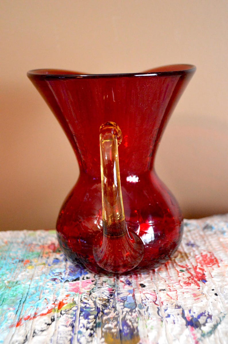 May include: A red glass pitcher with a clear handle. The pitcher has a bulbous base and a flared top. The handle is attached to the top and base of the pitcher. The pitcher is sitting on a multi-colored surface.