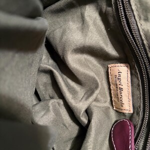 May include: Close-up of the interior of a green handbag. The bag features a zipper, a leather tag with the text "Angel Barcelo Designed in the EU", and a burgundy leather pull tab.