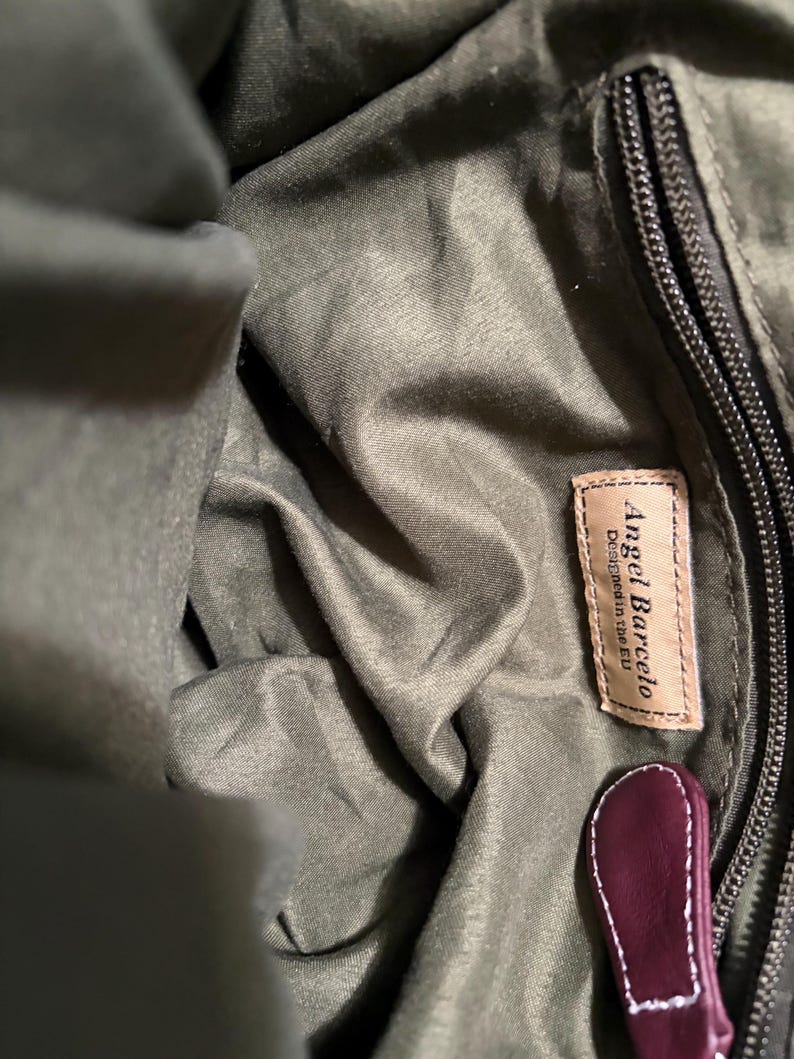 May include: Close-up of the interior of a green handbag. The bag features a zipper, a leather tag with the text "Angel Barcelo Designed in the EU", and a burgundy leather pull tab.