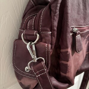 May include: A burgundy leather shoulder bag with white stitching. The bag features a zippered main compartment, a small front pocket, and a silver-tone metal clasp and hardware. The strap is adjustable.