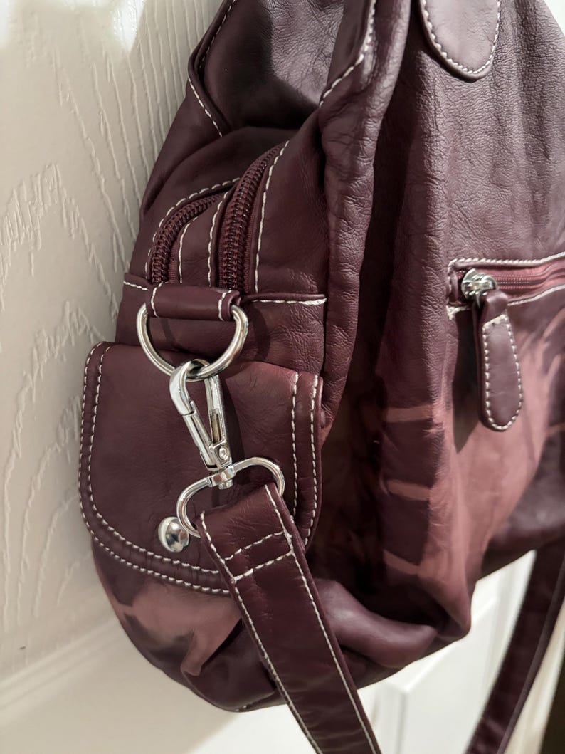 May include: A burgundy leather shoulder bag with white stitching. The bag features a zippered main compartment, a small front pocket, and a silver-tone metal clasp and hardware. The strap is adjustable.