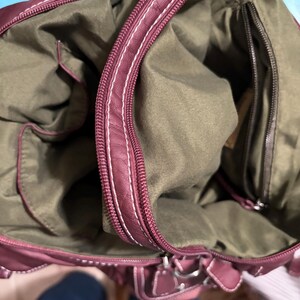 May include: Burgundy handbag with white stitching and a zipper closure. The interior is olive green with multiple pockets. The bag has a structured shape and a top handle.
