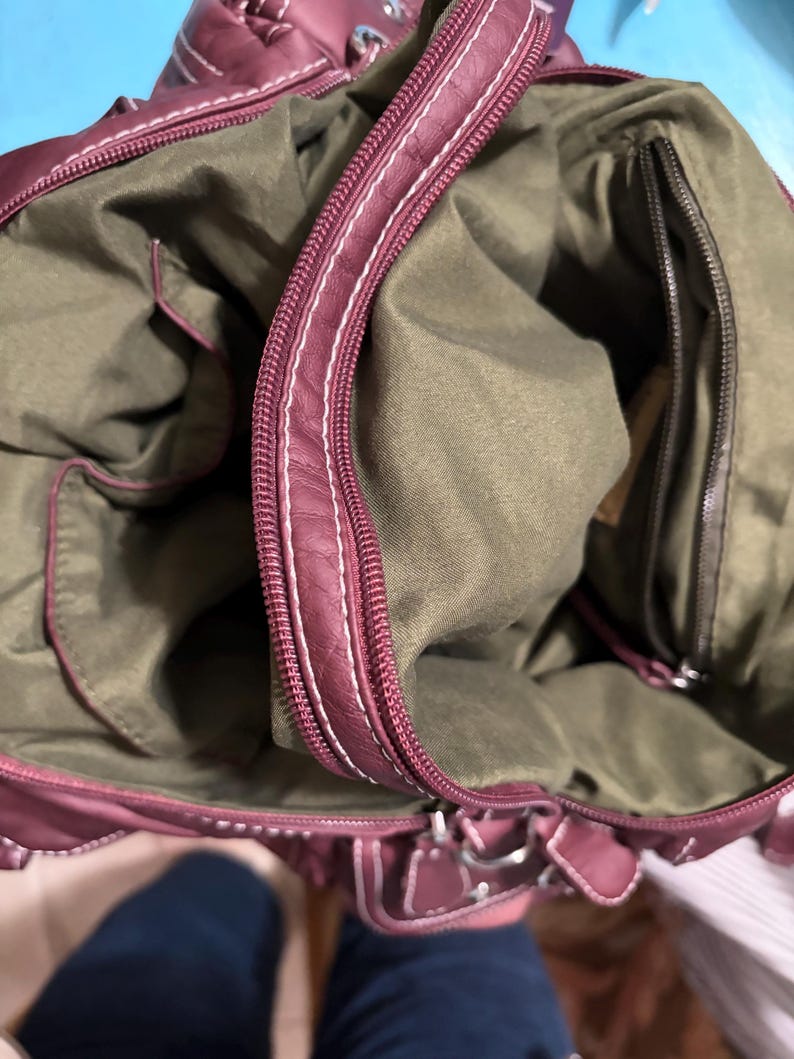 May include: Burgundy handbag with white stitching and a zipper closure. The interior is olive green with multiple pockets. The bag has a structured shape and a top handle.