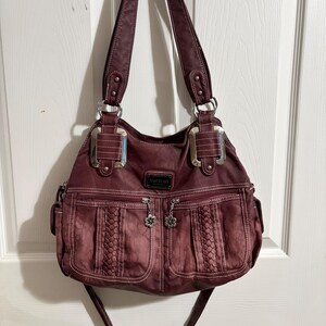 May include: A burgundy handbag with two top handles and a detachable shoulder strap. The bag features two front pockets with braided detailing and flower-shaped zipper pulls. Silver-tone hardware accents the bag's design.
