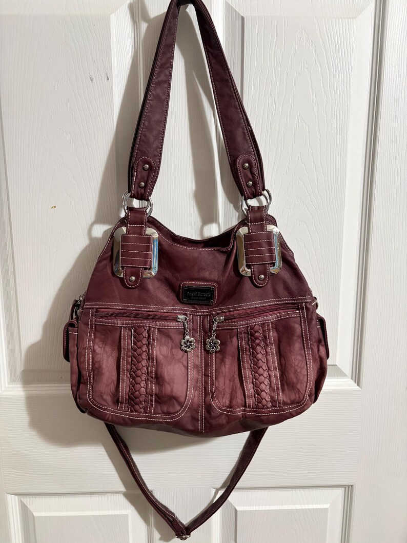 May include: A burgundy handbag with two top handles and a detachable shoulder strap. The bag features two front pockets with braided detailing and flower-shaped zipper pulls. Silver-tone hardware accents the bag's design.