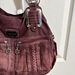 May include: A burgundy shoulder bag with white stitching and silver hardware. The bag features a braided design, two zippered pockets with flower charms, and the brand name "Angel Barcelo". The bag has a textured appearance.