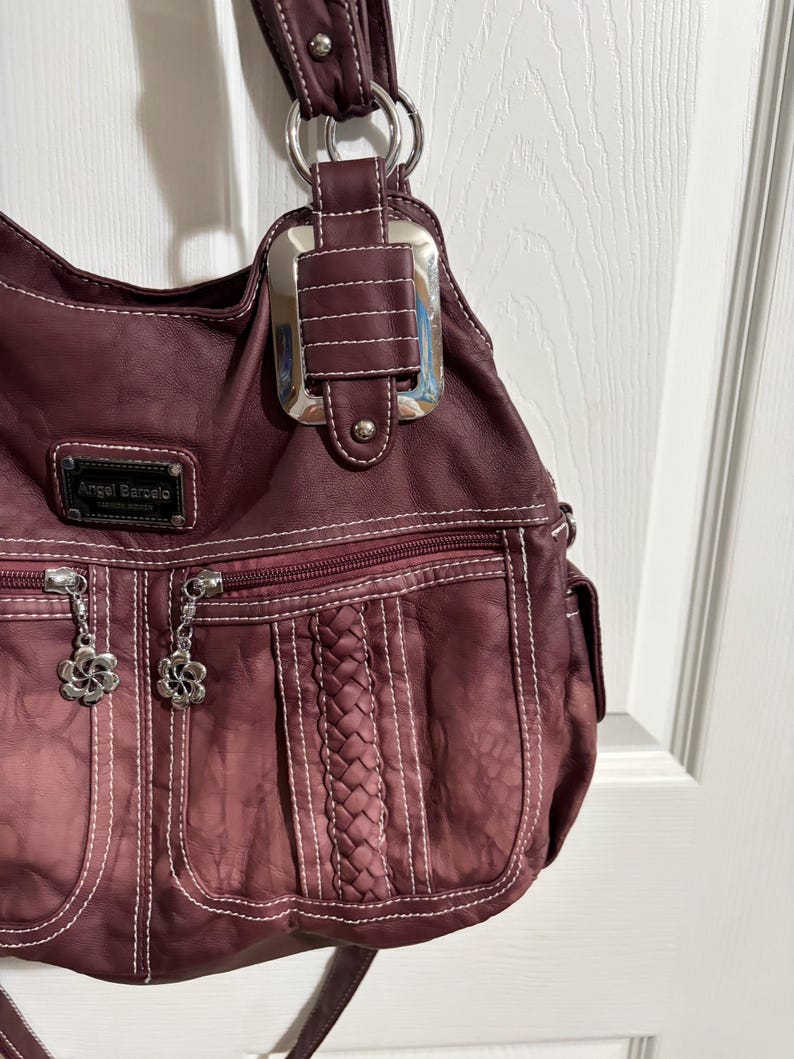 May include: A burgundy shoulder bag with white stitching and silver hardware. The bag features a braided design, two zippered pockets with flower charms, and the brand name "Angel Barcelo". The bag has a textured appearance.