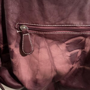 May include: A close-up of a burgundy leather bag with a zipper pocket. The bag has white stitching and a matching leather zipper pull. The leather has a faded, textured appearance.