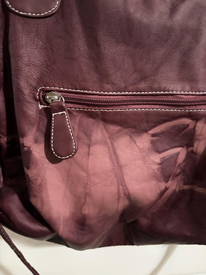 May include: A close-up of a burgundy leather bag with a zipper pocket. The bag has white stitching and a matching leather zipper pull. The leather has a faded, textured appearance.