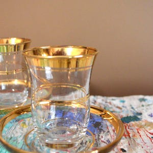 May include: Clear glass teacups with gold rims and gold accent lines, resting on matching gold-rimmed saucers. The teacups are on a colorful, patterned surface, with a neutral background.