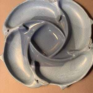 May include: A light blue ceramic serving dish with a unique design. The dish features a central square compartment surrounded by three curved sections, each with a dolphin-like shape. The speckled finish adds texture to the piece.