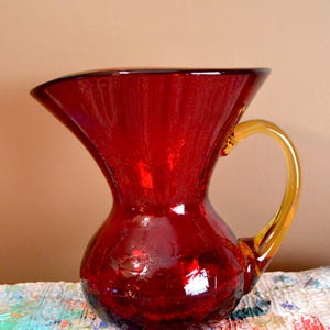 May include: A vibrant red glass pitcher with a flared top and a rounded base. The pitcher features a contrasting amber-colored handle. This decorative piece is ideal for serving beverages or as a decorative accent.