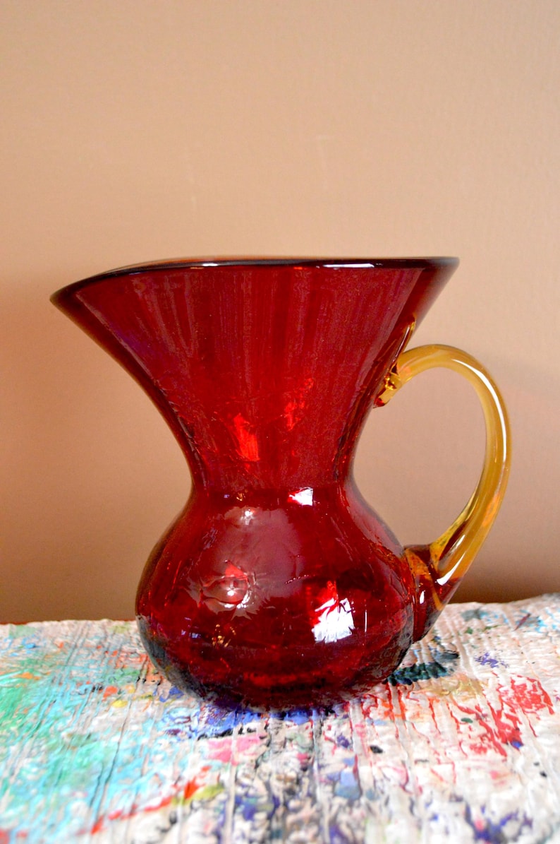 May include: A vibrant red glass pitcher with a flared top and a rounded base. The pitcher features a contrasting amber-colored handle. This decorative piece is ideal for serving beverages or as a decorative accent.