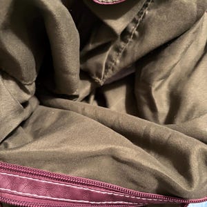 May include: Interior view of a burgundy handbag with a dark olive green lining. The bag's zipper is burgundy with white stitching. The lining is a soft, textured fabric.