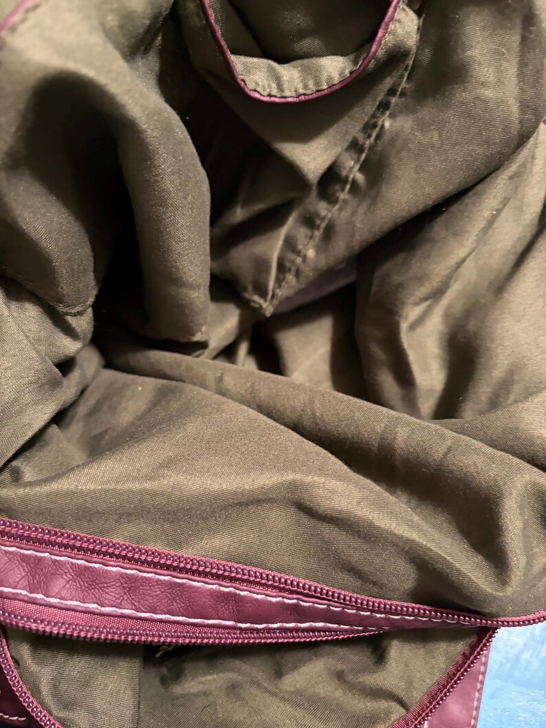 May include: Interior view of a burgundy handbag with a dark olive green lining. The bag's zipper is burgundy with white stitching. The lining is a soft, textured fabric.