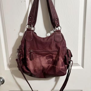May include: A burgundy shoulder bag with a tie-dye effect. The bag has two top handles and a long, adjustable strap. Features include a zippered main compartment, exterior pockets, and silver-tone hardware. White stitching accents the bag's seams.