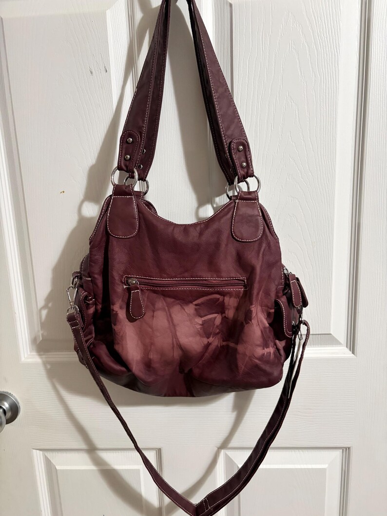May include: A burgundy shoulder bag with a tie-dye effect. The bag has two top handles and a long, adjustable strap. Features include a zippered main compartment, exterior pockets, and silver-tone hardware. White stitching accents the bag's seams.