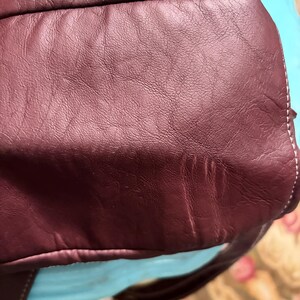 May include: Close-up of a burgundy leather bag. The bag has a textured surface with visible grain and subtle variations in color. White stitching is visible along the edges. The bag appears to be well-worn and has a soft, supple appearance.