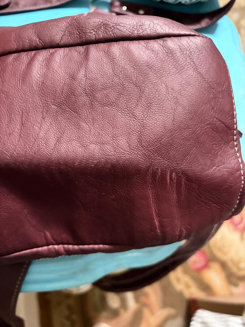 May include: Close-up of a burgundy leather bag. The bag has a textured surface with visible grain and subtle variations in color. White stitching is visible along the edges. The bag appears to be well-worn and has a soft, supple appearance.
