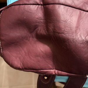 May include: A burgundy leather handbag with white stitching. The bag has a rounded shape and a handle. The leather appears to be textured. The bag is a deep shade of red-purple.
