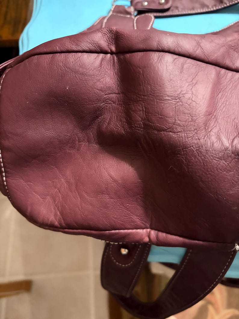 May include: A burgundy leather handbag with white stitching. The bag has a rounded shape and a handle. The leather appears to be textured. The bag is a deep shade of red-purple.