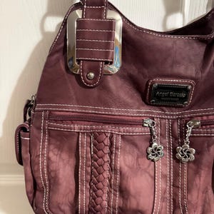 May include: A burgundy hobo-style handbag with white stitching and silver-tone hardware. Features include a braided accent, multiple zippered pockets, and flower-shaped zipper pulls. The brand name "Angel Barcelo" is visible on a small tag.