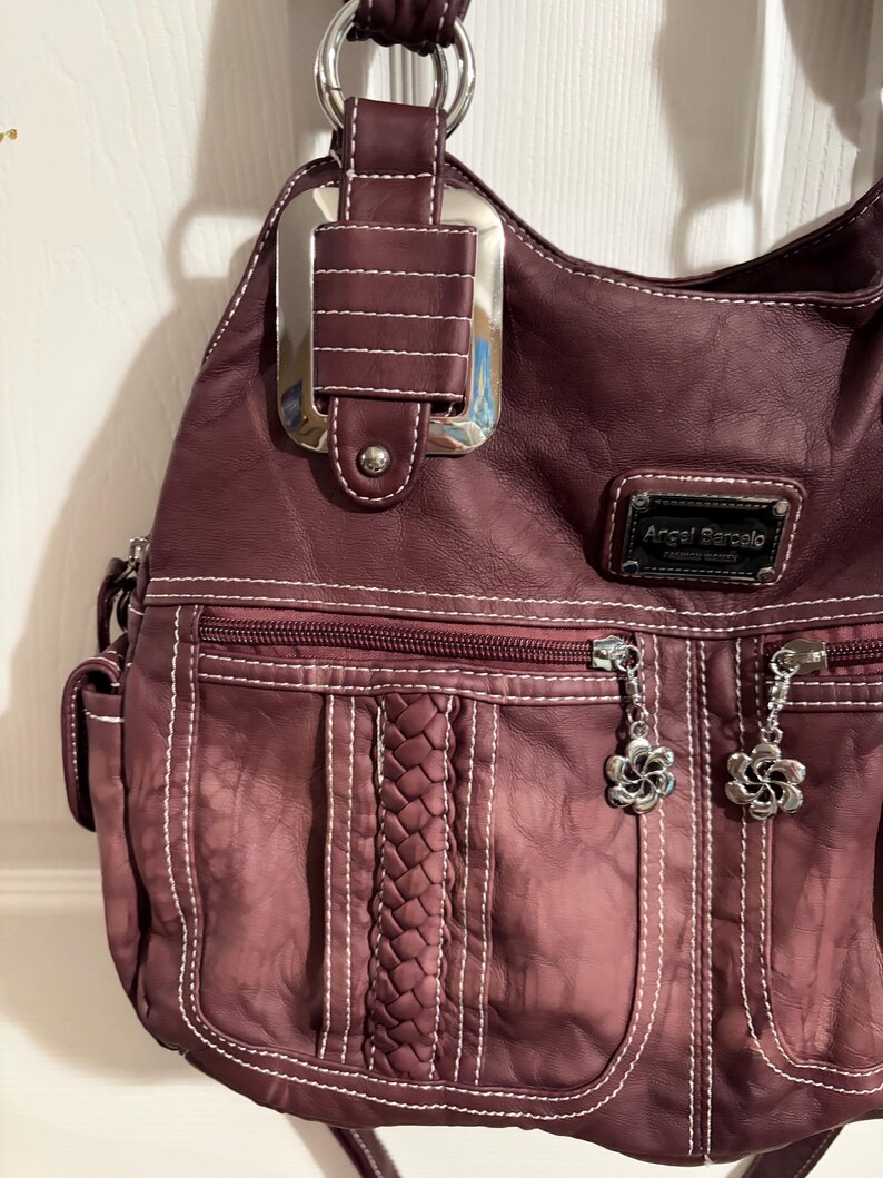 May include: A burgundy hobo-style handbag with white stitching and silver-tone hardware. Features include a braided accent, multiple zippered pockets, and flower-shaped zipper pulls. The brand name "Angel Barcelo" is visible on a small tag.