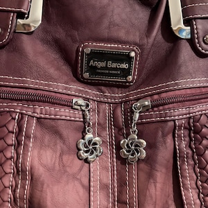 May include: A burgundy leather handbag with silver hardware and a zipper closure. The bag features white stitching, a braided texture, and two flower-shaped zipper pulls. The label reads "Angel Barcelo FASHION WOMEN."