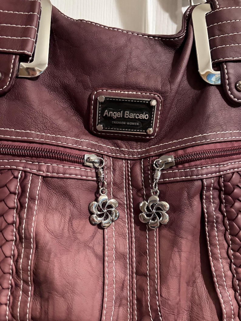 May include: A burgundy leather handbag with silver hardware and a zipper closure. The bag features white stitching, a braided texture, and two flower-shaped zipper pulls. The label reads "Angel Barcelo FASHION WOMEN."