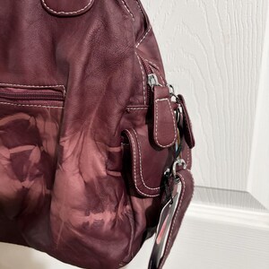 May include: A burgundy leather handbag with white stitching. The bag features multiple pockets, a zipper closure, and a shoulder strap. The leather has a unique, faded pattern. A tag hangs from the strap.