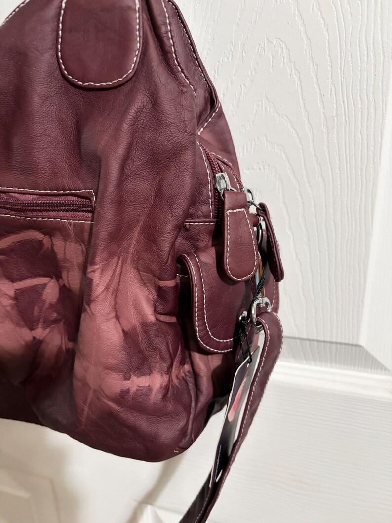 May include: A burgundy leather handbag with white stitching. The bag features multiple pockets, a zipper closure, and a shoulder strap. The leather has a unique, faded pattern. A tag hangs from the strap.