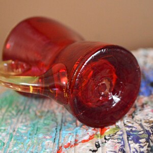 May include: A red glass pitcher with a clear handle. The pitcher is lying on its side, revealing its base. The handle is a lighter shade of red and appears to be made of the same material. The pitcher is on a surface with colorful paint splatters.