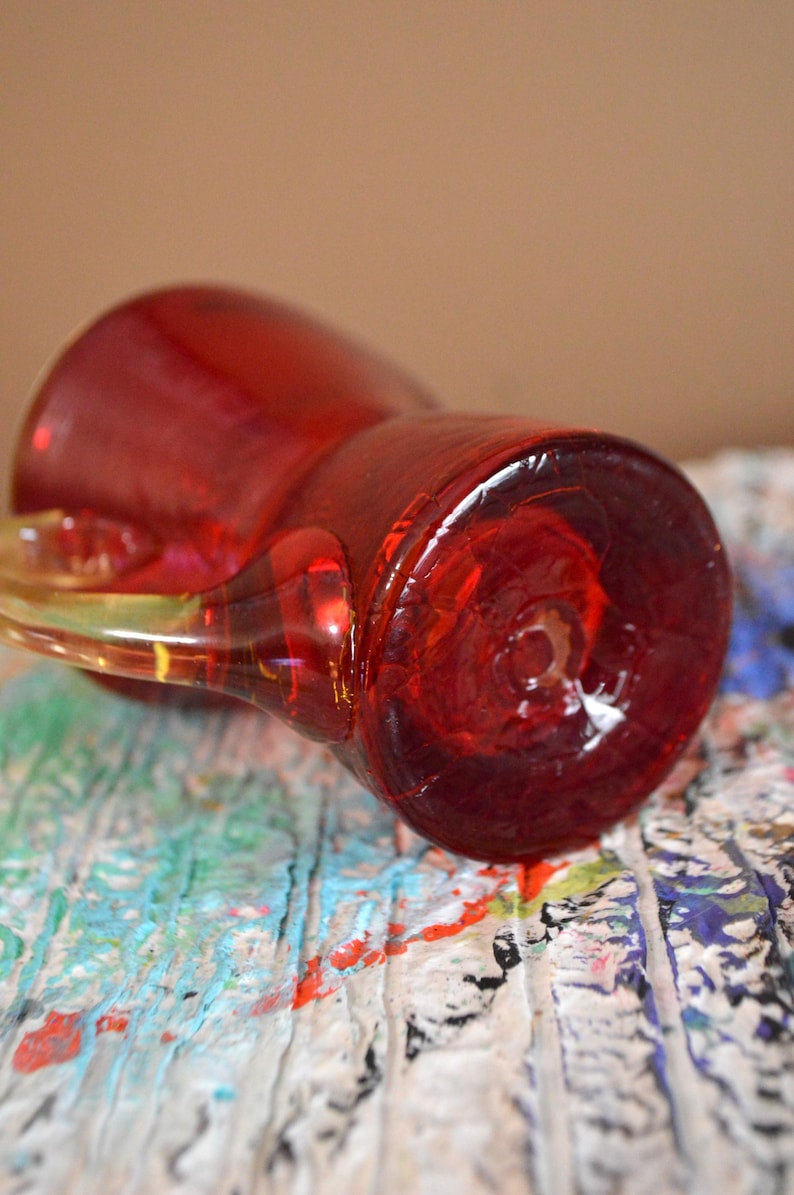 May include: A red glass pitcher with a clear handle. The pitcher is lying on its side, revealing its base. The handle is a lighter shade of red and appears to be made of the same material. The pitcher is on a surface with colorful paint splatters.