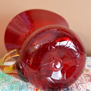 May include: A handcrafted, red glass mug with a rounded body and a clear handle. The mug is positioned on a colorful, textured surface, with a matching red saucer in the background. The glass has a crackled appearance.