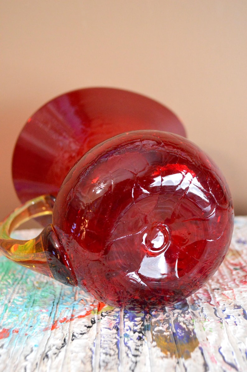 May include: A handcrafted, red glass mug with a rounded body and a clear handle. The mug is positioned on a colorful, textured surface, with a matching red saucer in the background. The glass has a crackled appearance.