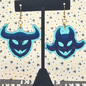 May include: A pair of Halloween-themed earrings. Each earring features a dark blue devil face design with a light blue outline. The earrings are hanging from gold-colored hooks. The background is a patterned paper with spiders.