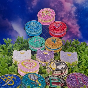 Könnte beinhalten: Eine Sammlung von 12 bunten Emaille-Pins in Form von Macarons mit verschiedenen Designs, darunter ein Stern, ein Halbmond, ein Herz und ein Schwert. Die Pins sind in Pyramidenform auf einem Bett aus grünen Blättern angeordnet.