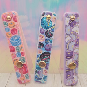 Puede incluir: Tres agarres para teléfono con diferentes patrones. El primer agarre es rosa con un patrón de macarons coloridos. El segundo agarre es azul con un patrón de macarons azules y negros. El tercer agarre es morado con un patrón de nubes y estrellas blancas y moradas.