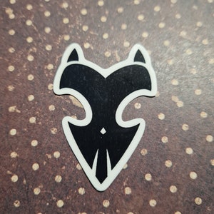 May include: A black and white sticker with a stylized fox head design. The sticker features a black fox head shape with pointed ears and a white outline. The design is symmetrical and has a minimalist aesthetic.