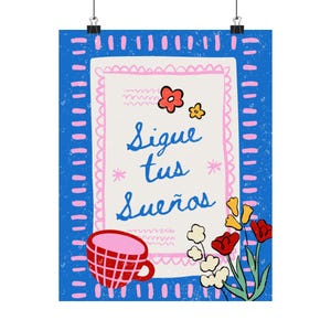 May include: A rectangular wall poster with a blue background featuring a pink dashed border. The center contains the text Sigue tus sueños in blue script, surrounded by small red and yellow flowers, and a red checkered cup at the bottom.
